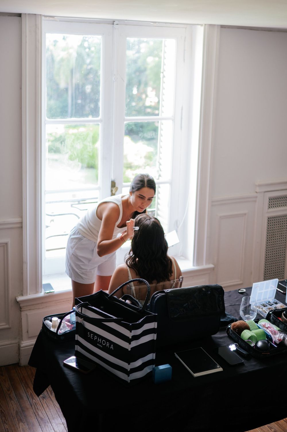Makeup artist applying cosmetics to bride near window with Sephora bag on table.