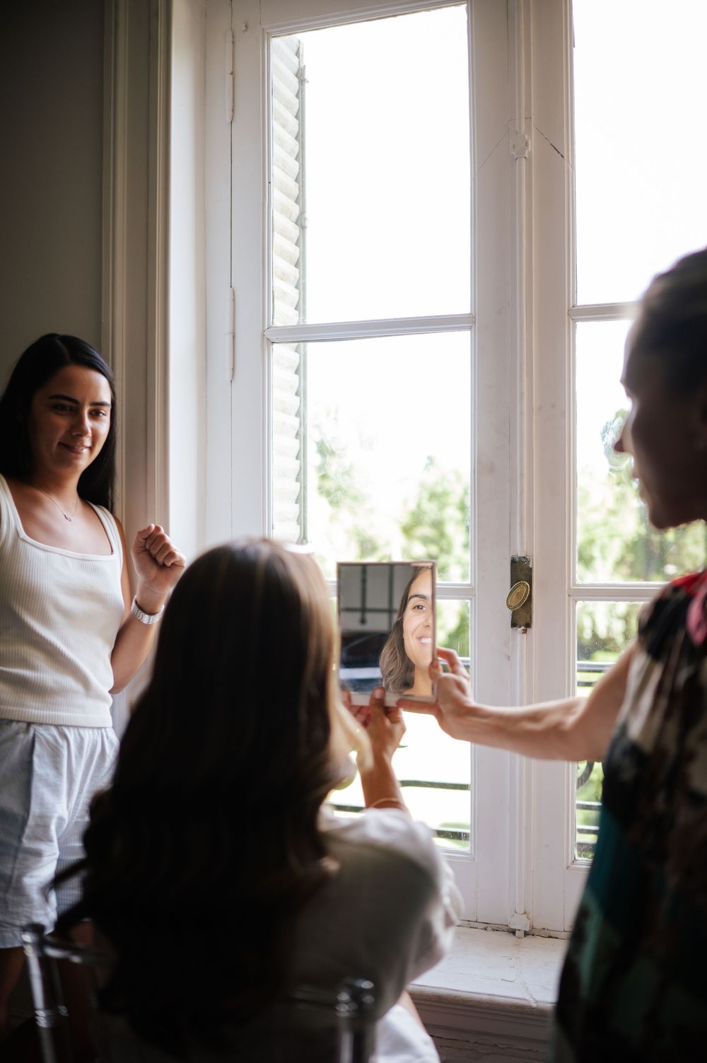 Bride admiring her makeup in mirror held by stylist near bright window.
