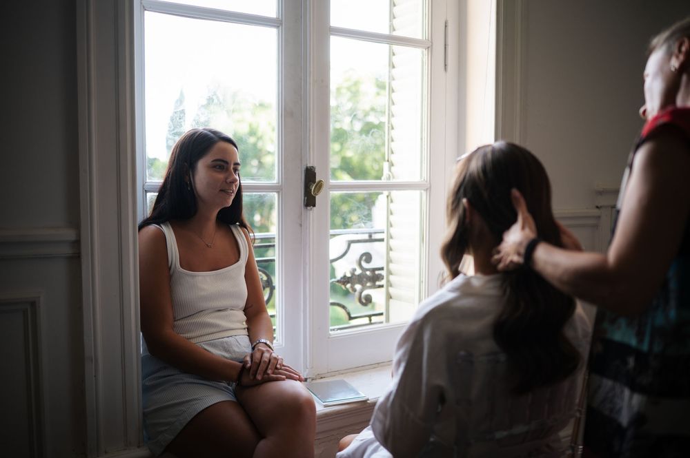 Bride sitting by window while hairstylist works on her hair during prep.