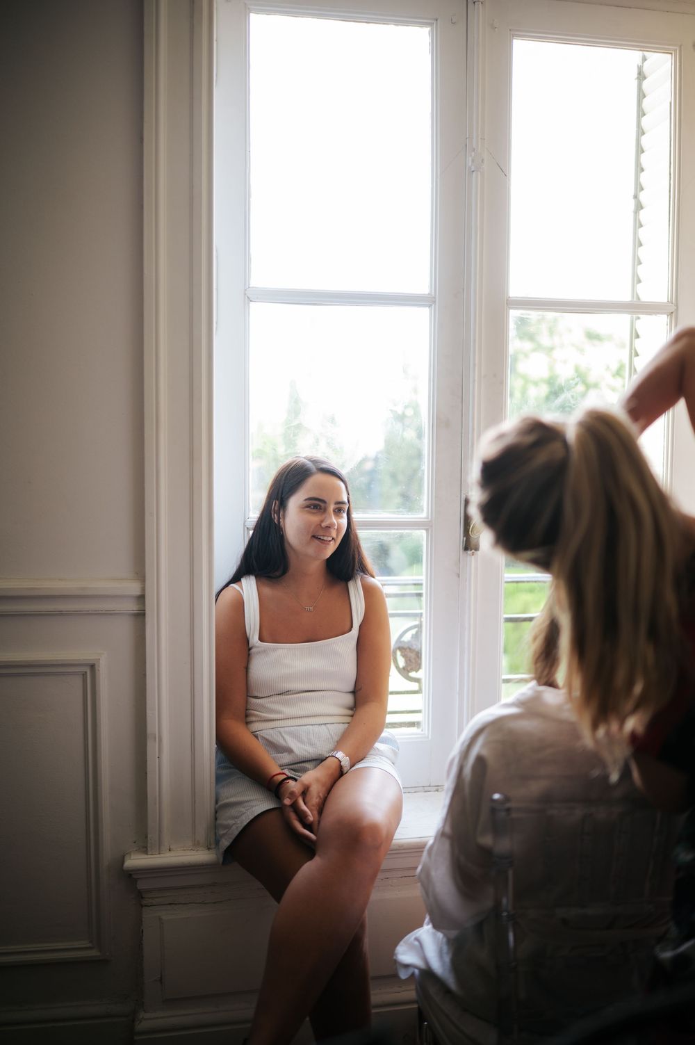 Smiling bride sitting on windowsill during wedding morning hair preparation.