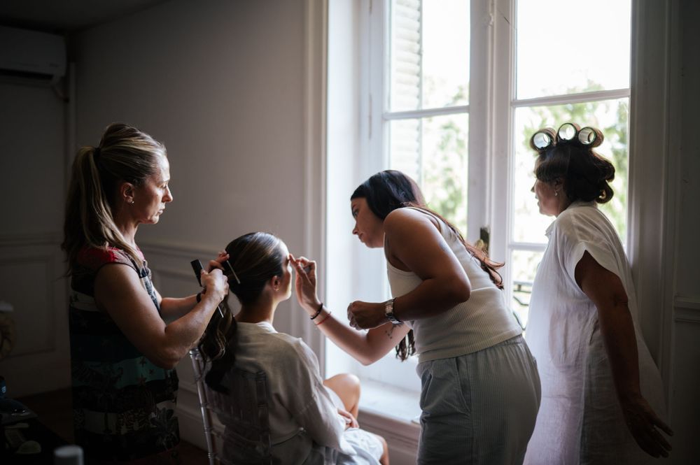 Bridesmaids getting hair styled by professionals near bright windows before a wedding ceremony.