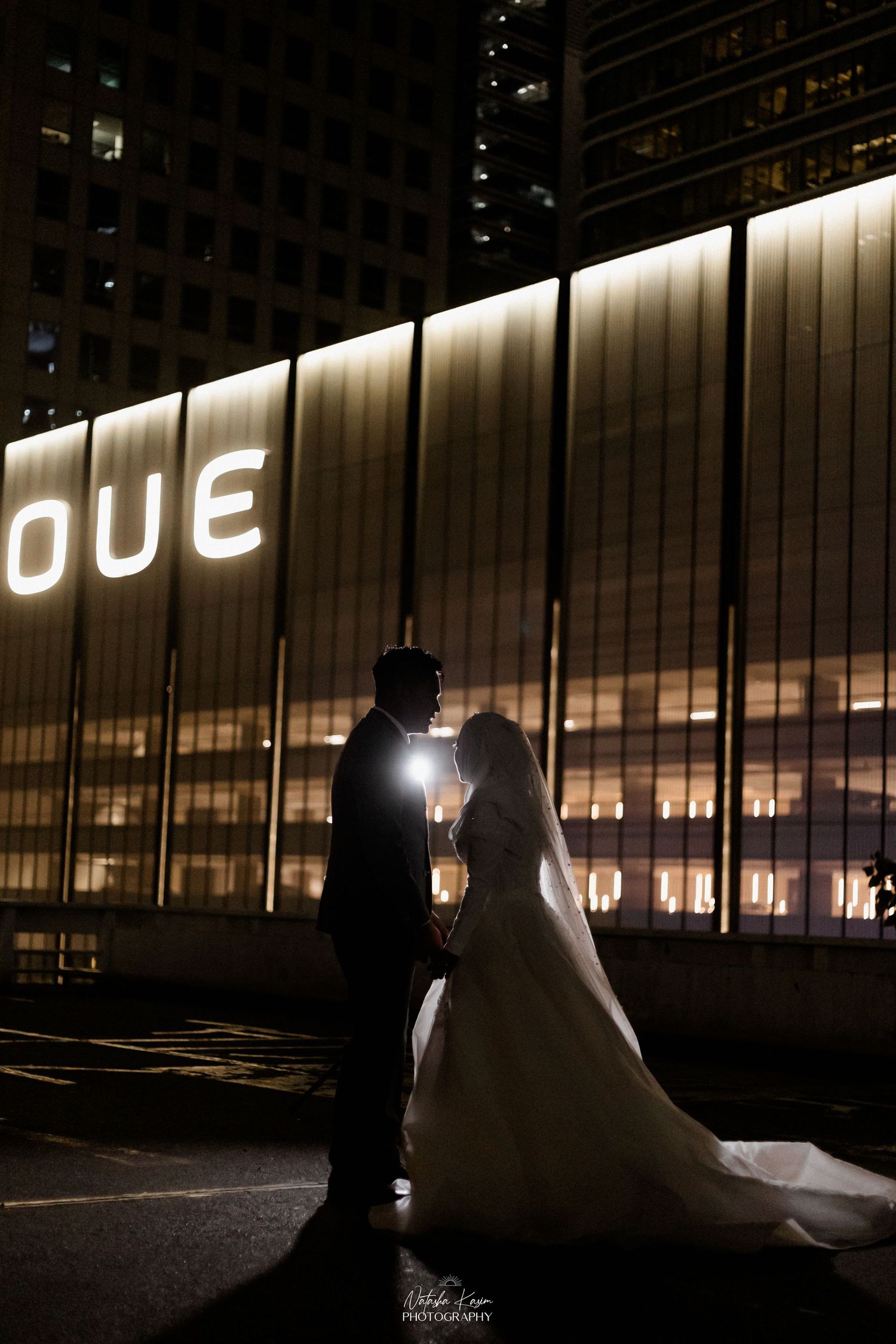 Muslim couple walking hand in hand in a lush garden, woman in hijab and white dress, man in cream jacket.