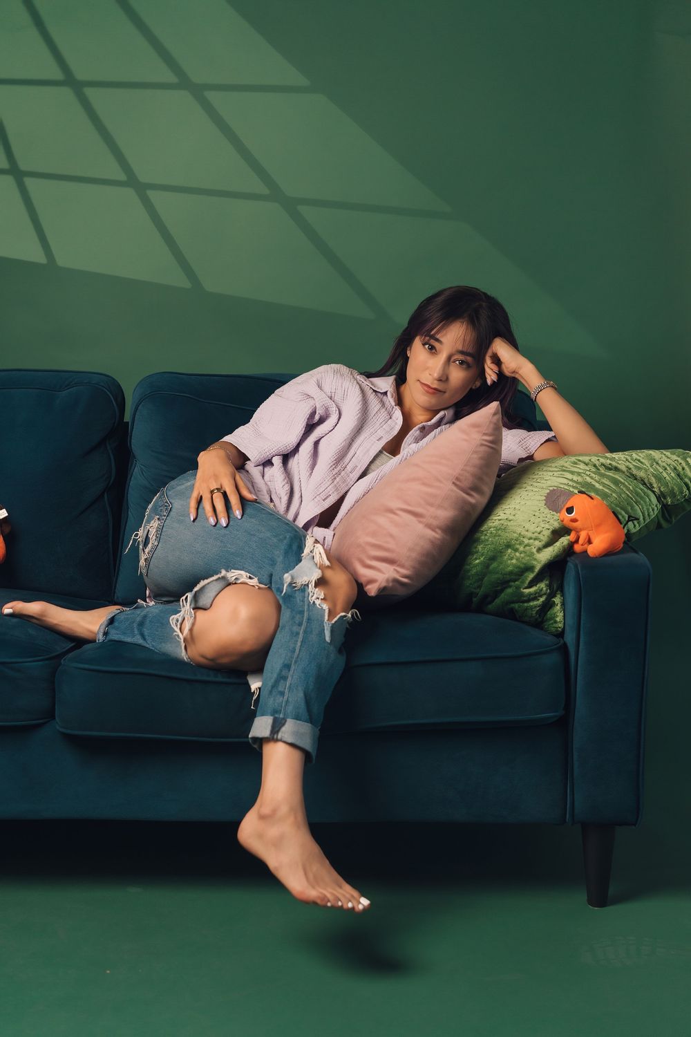 Woman in ripped jeans and lilac shirt relaxing on teal velvet sofa with colorful pillows against green wall.