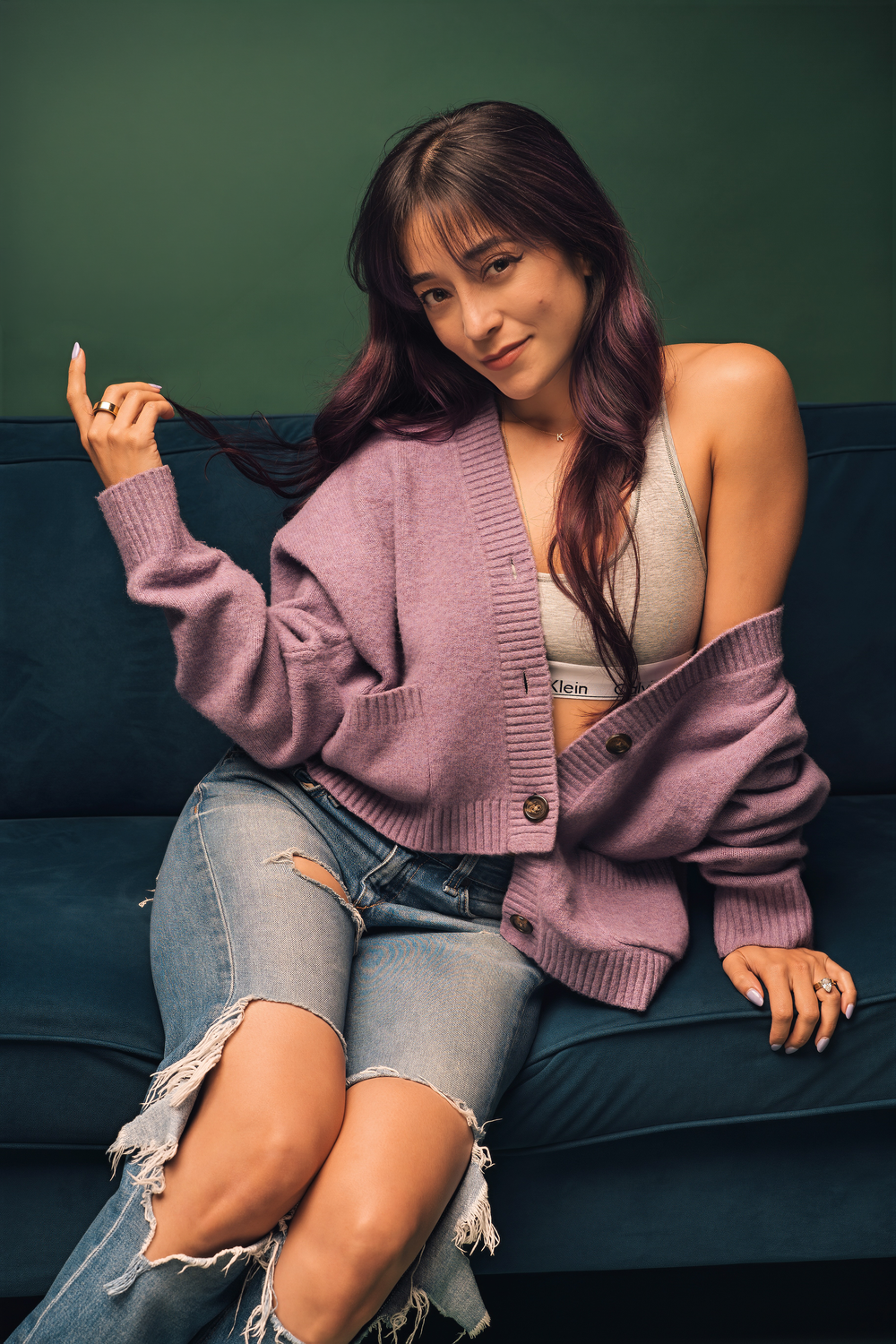 Young woman in pink cardigan and ripped jeans sitting relaxed on a teal sofa, smiling softly.
