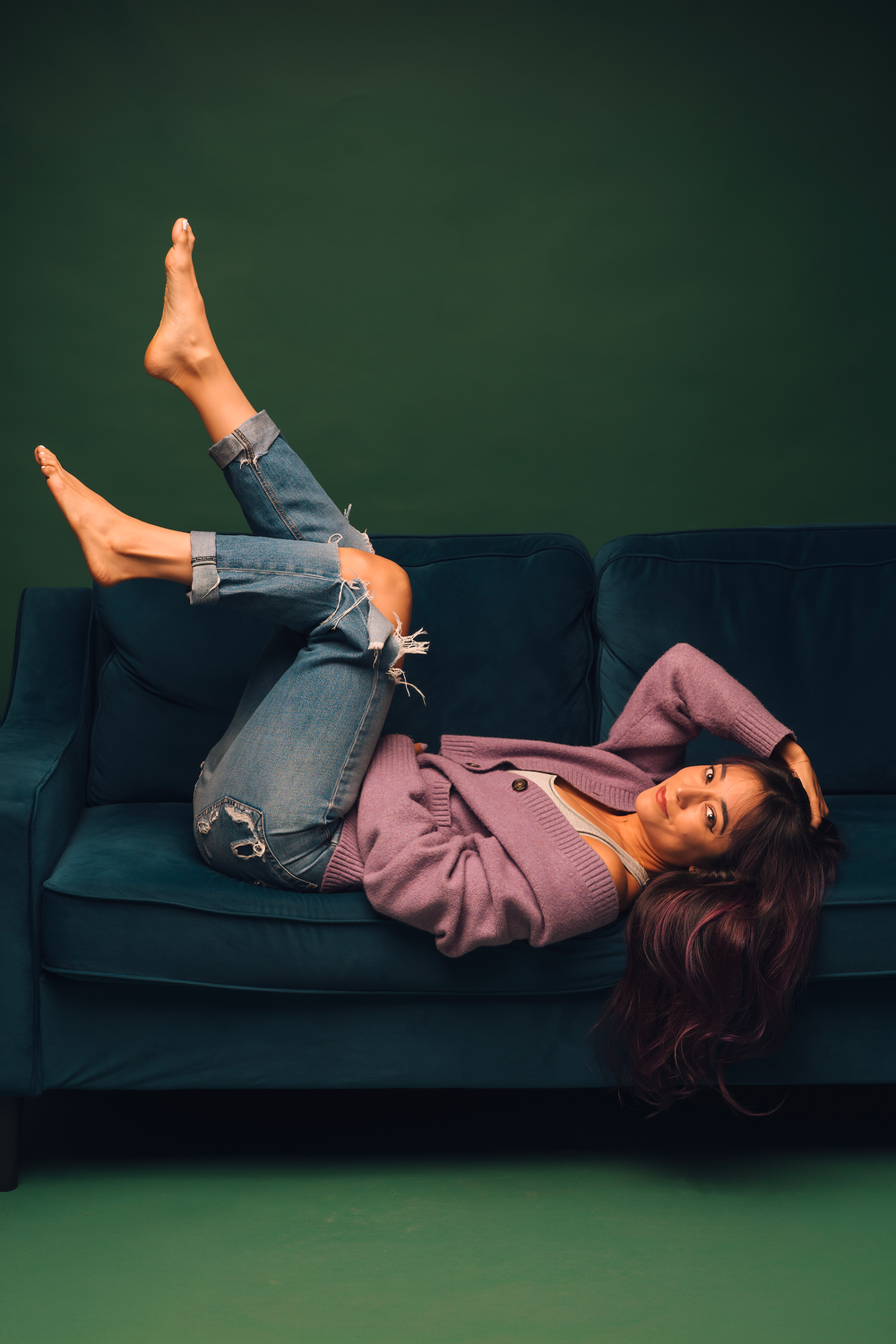 Woman in ripped jeans and pink blazer lying on a teal velvet sofa with legs raised, against a dark green background.