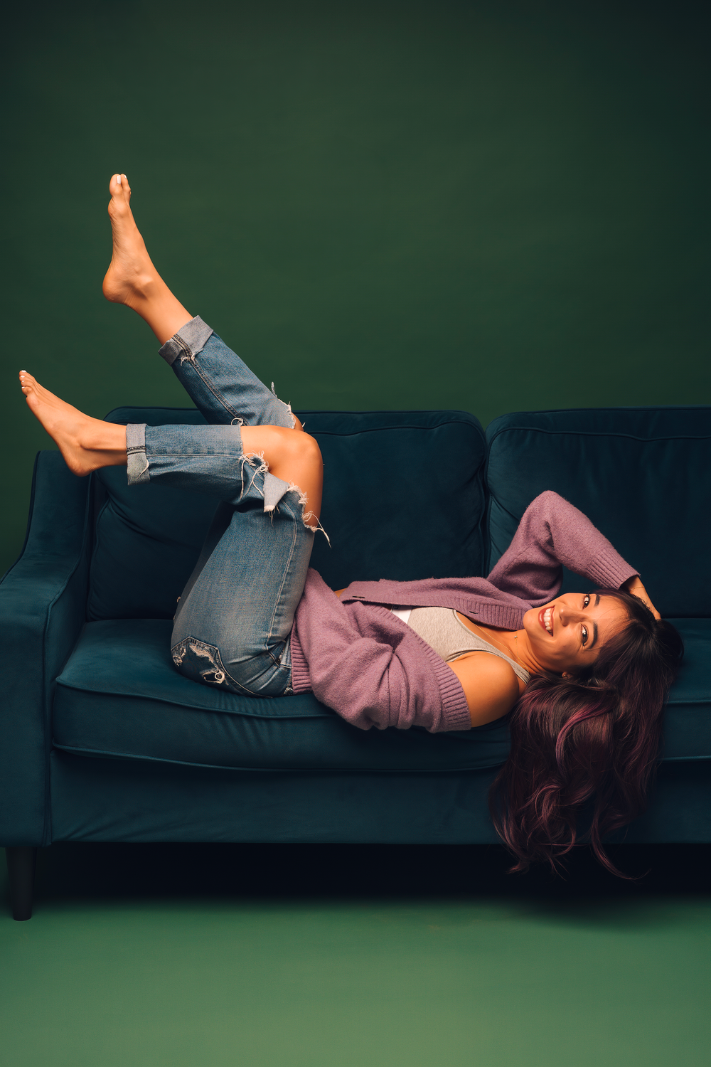 Woman in ripped jeans and purple cardigan relaxing on a teal velvet sofa against a dark green background.