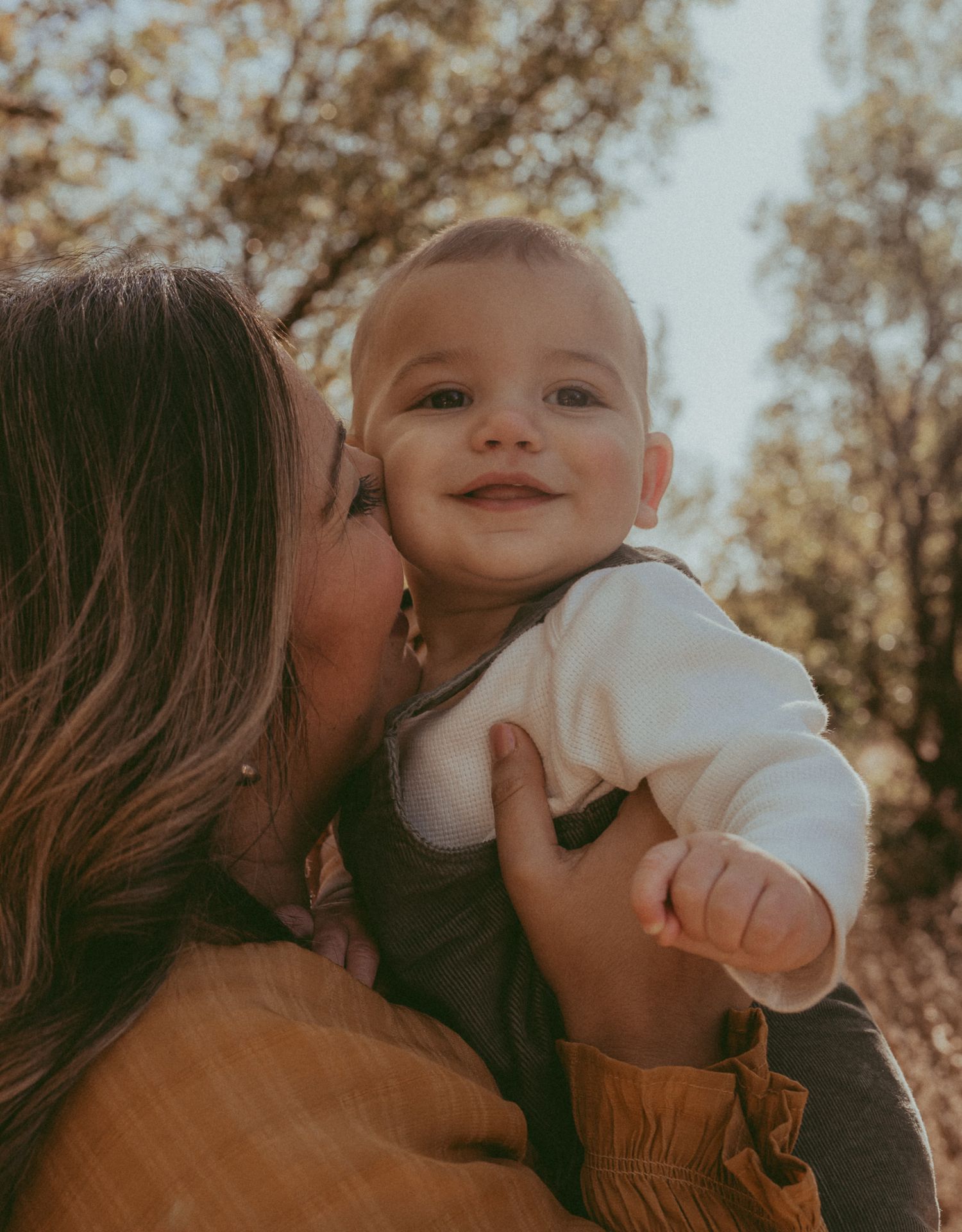 A mother lovingly kisses her smiling baby outdoors in warm golden light among autumn trees.