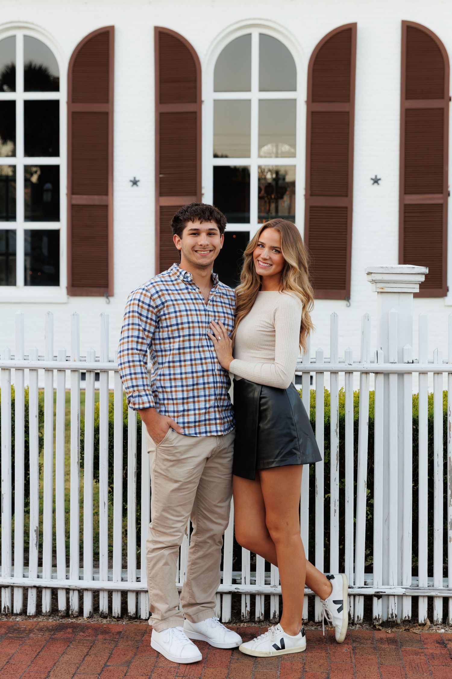 A young couple smiling together in front of a white picket fence and building with brown shutters.