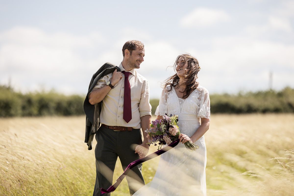 A bride and groom smile at each other in a sunny field, she holds a bouquet while he carries his jacket over his shoulder.