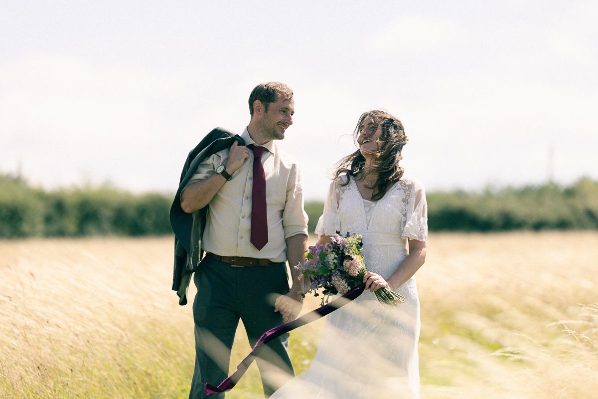 A smiling bride and groom walk through a sunny field, she holds a colorful bouquet in her white wedding dress.