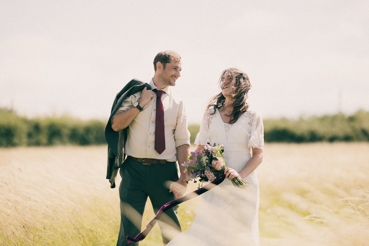 A bride and groom share a joyful moment in a sunny field, she holds a wildflower bouquet in her wedding dress.
