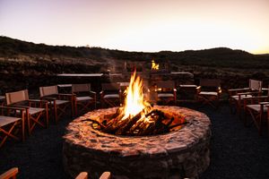 Stone fire pit with roaring flames surrounded by wooden chairs at dusk in a rugged safari landscape.