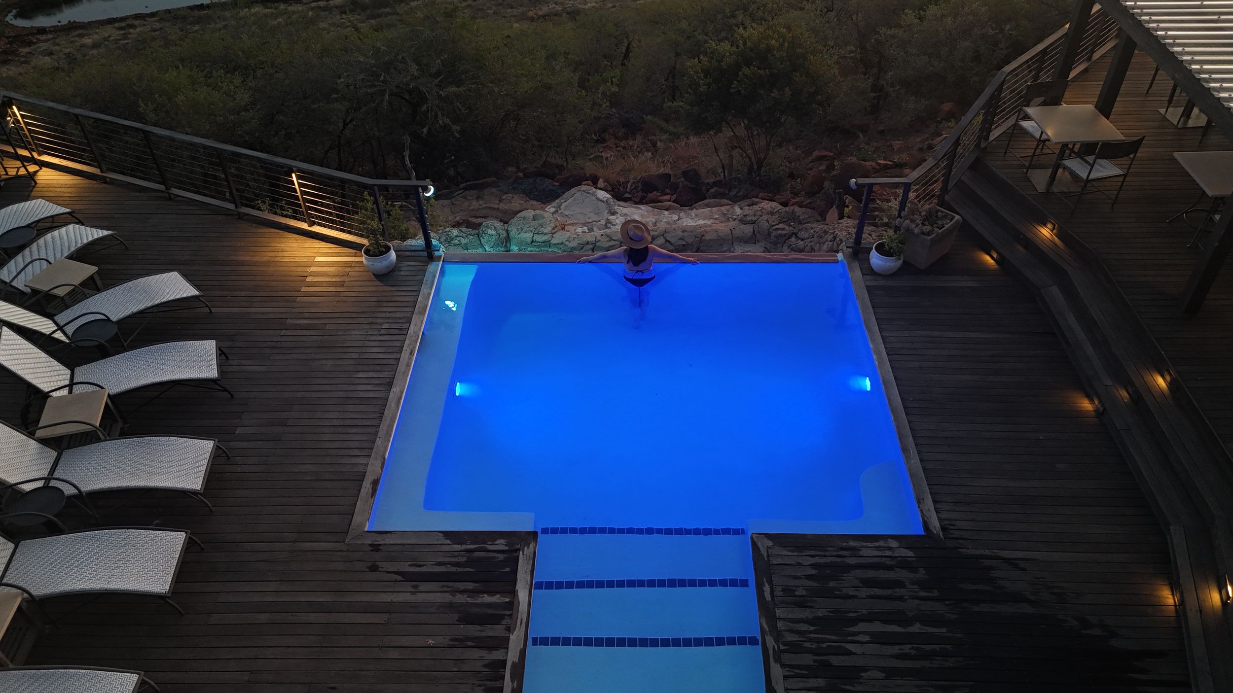 Aerial view of a glowing blue swimming pool on a wooden deck at dusk, surrounded by lounge chairs and natural rock at a Luxury Safari Lodge.