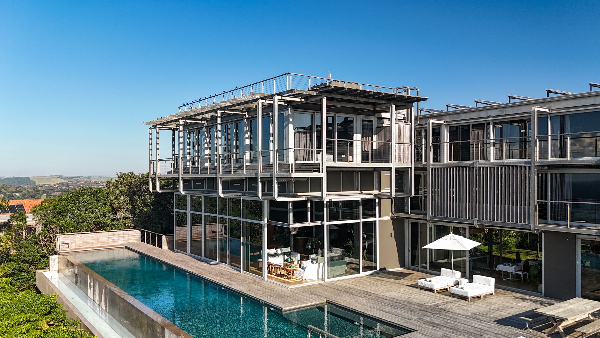 Modern multi-story glass and steel luxury villa with infinity pool, outdoor lounge chairs, and panoramic hillside views.