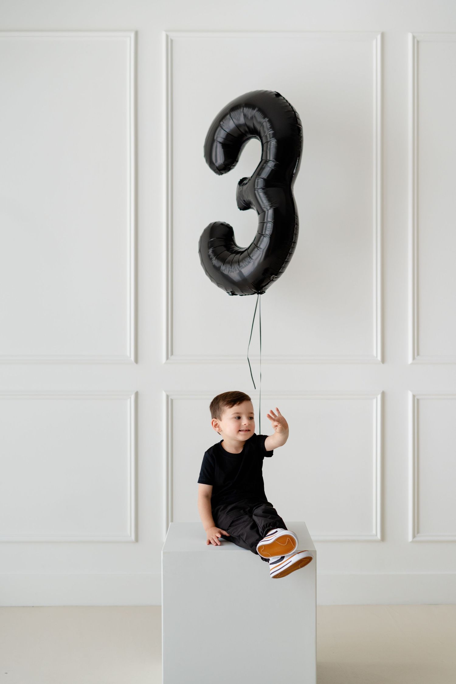 Toddler boy in black outfit sitting on white cube holding a large black number 3 balloon for his 3rd birthday.
