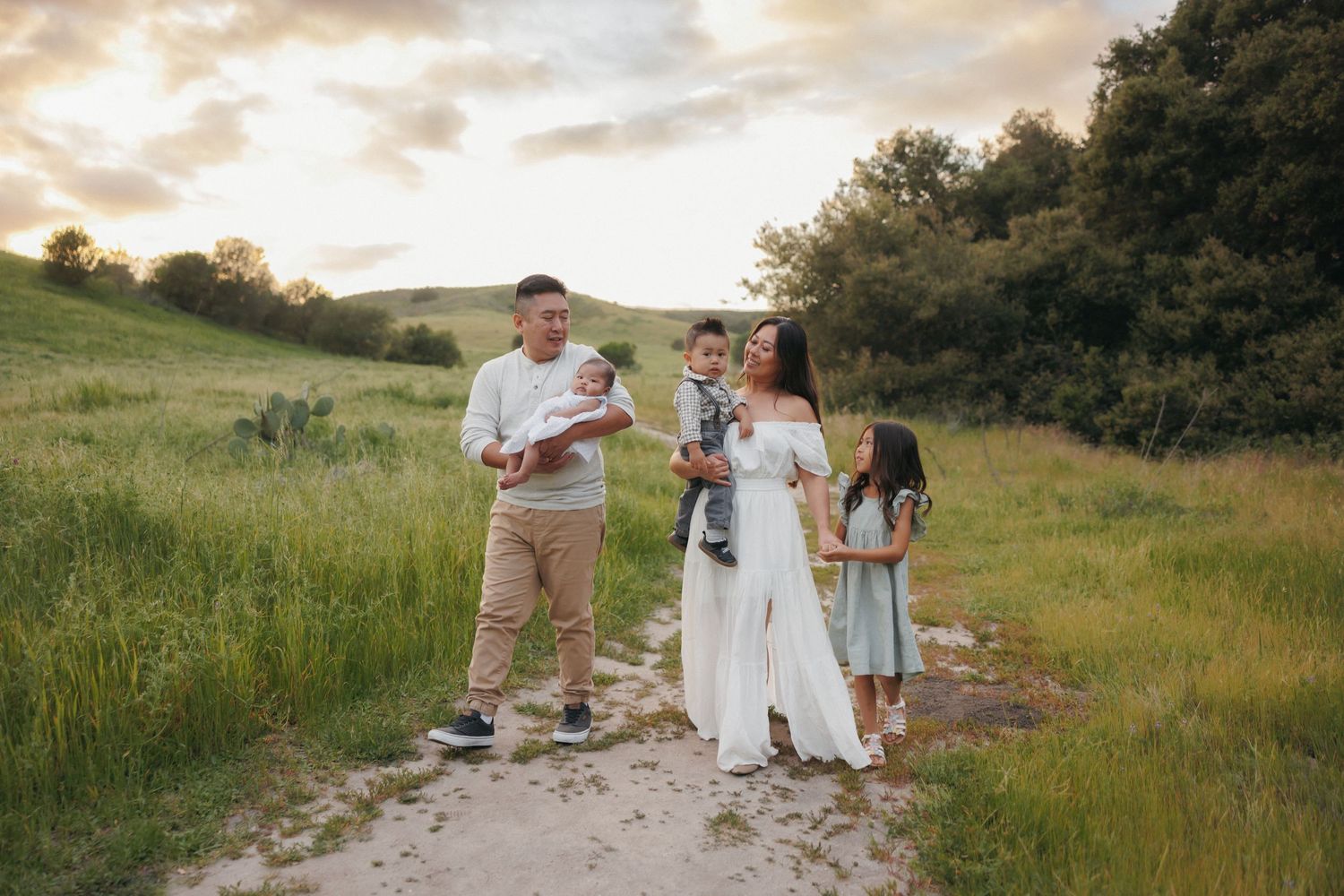 Family of five poses on a dirt path surrounded by lush green hills and trees during golden hour sunset.