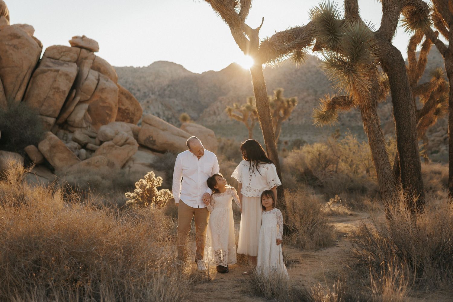 Family of four in white outfits walking through Joshua Tree desert landscape at golden sunset.