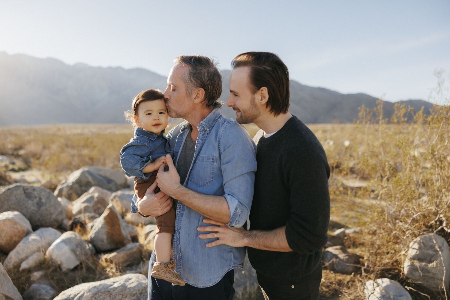 Two dads share a tender moment outdoors, one kissing their baby while the other embraces them against a mountain backdrop.