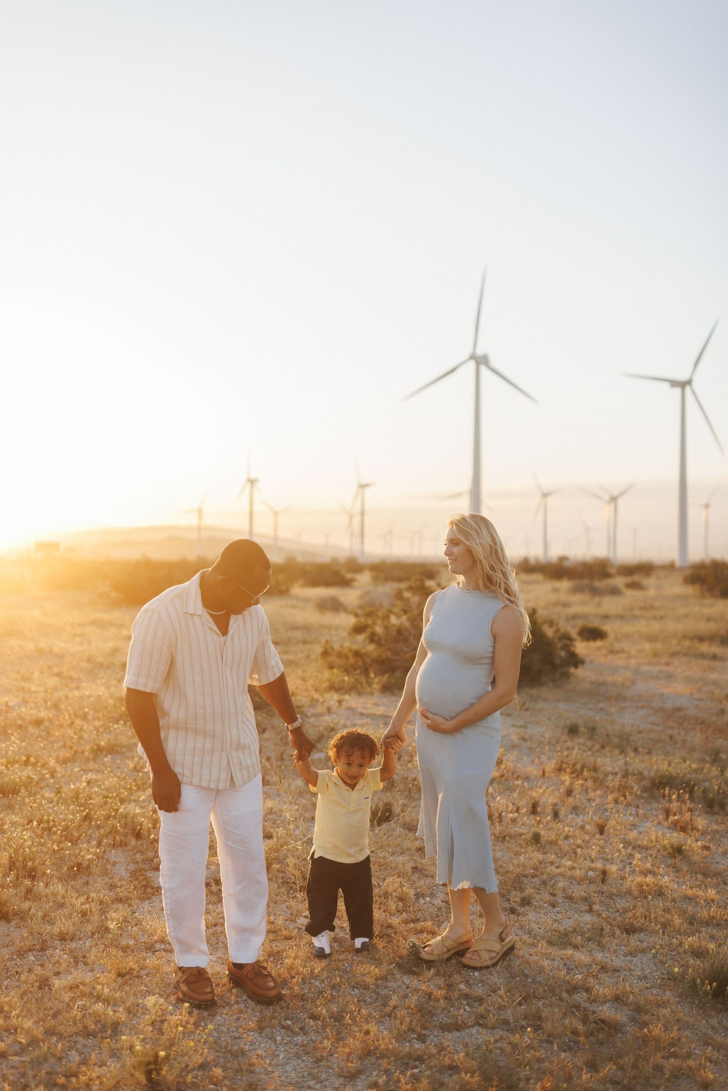 A pregnant woman and man hold hands with a toddler at sunset near wind turbines in a golden field.