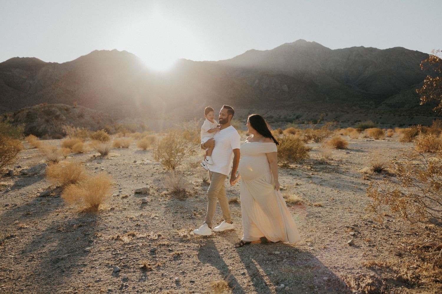 Couple in white outfits dancing in a sunlit desert landscape with mountains in the background.