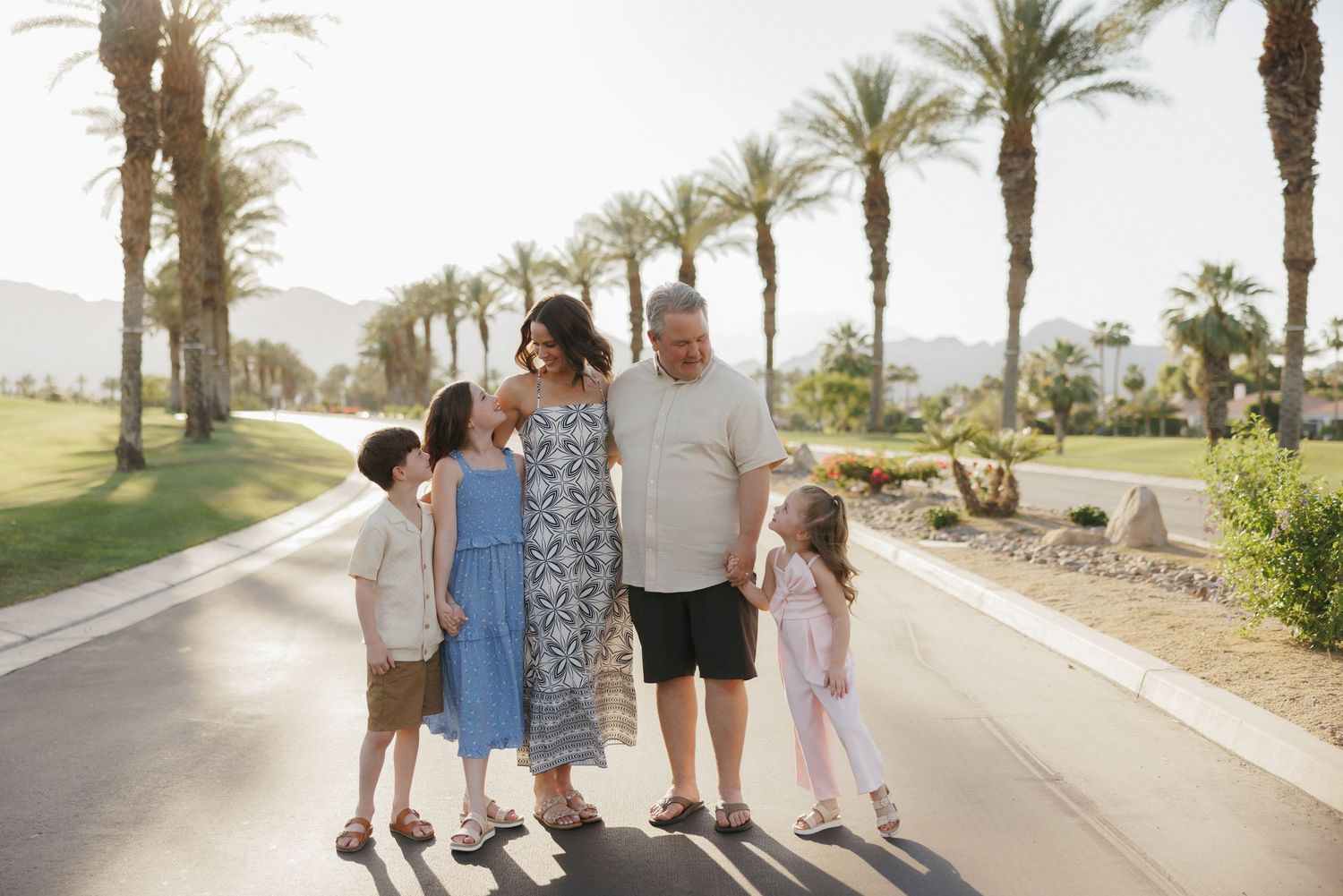 Family of five walking together on a sunny palm-lined path in a tropical resort setting.
