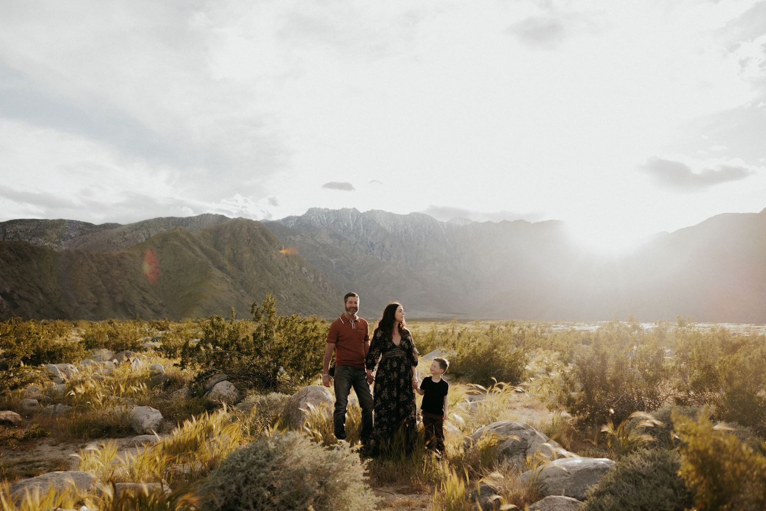 Family of three standing in desert landscape with mountain backdrop and golden sunlight, surrounded by cacti and shrubs.