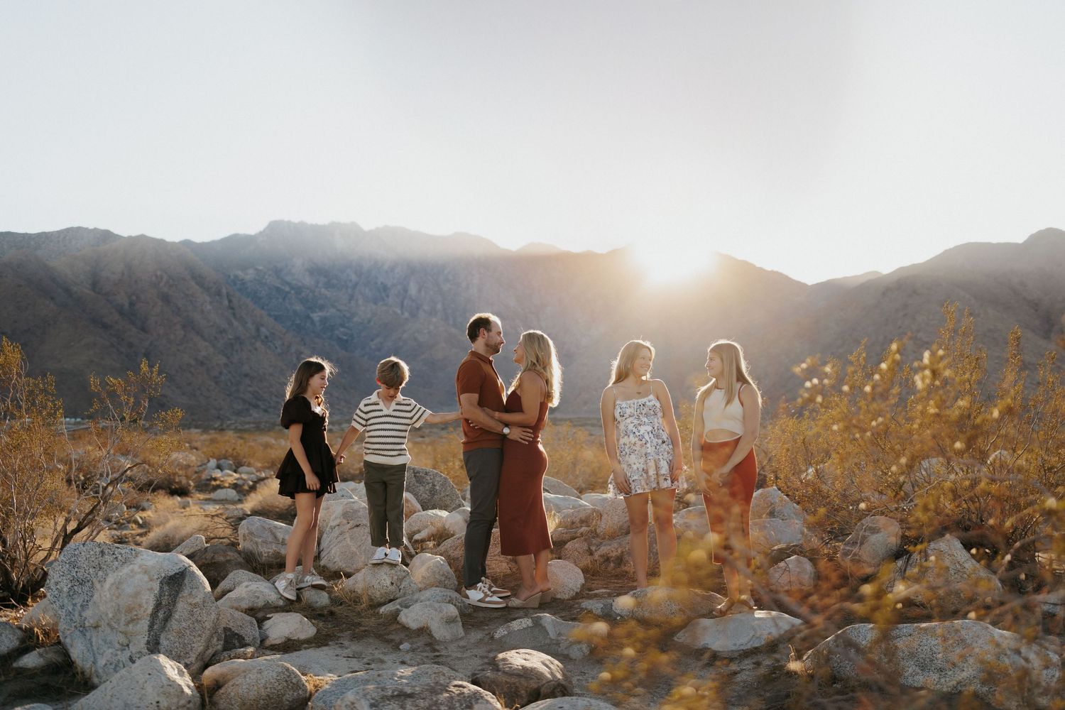 Family group standing on rocky desert terrain at golden hour with mountains and bright sunset in the background.