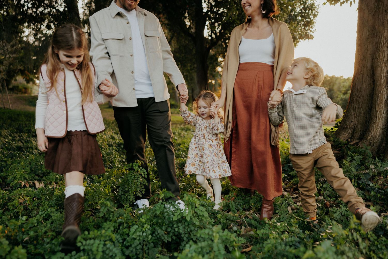 Family of five walking outdoors among green foliage, parents holding hands with three young children in warm autumn tones.