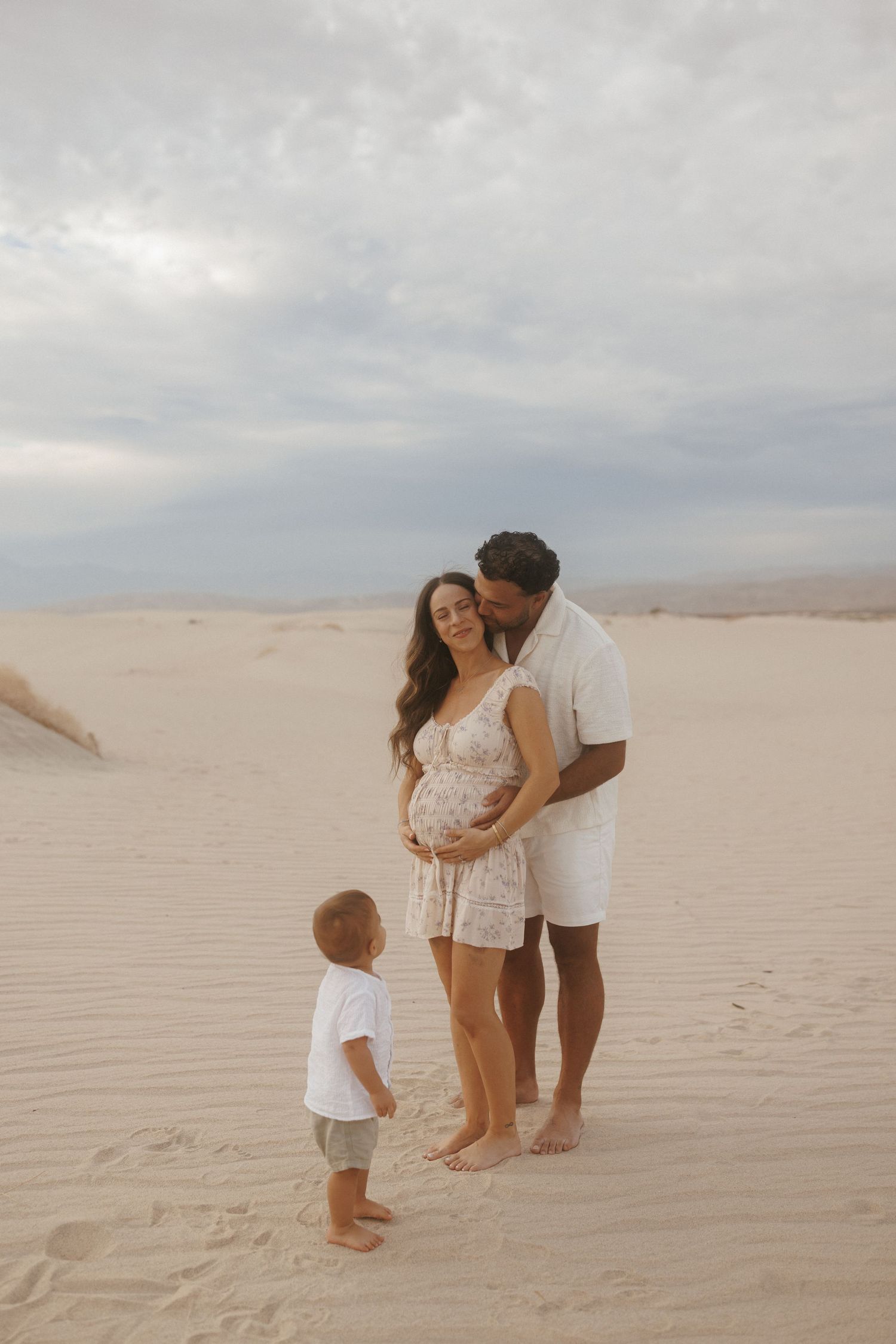 A pregnant woman and man embrace on sandy dunes while their toddler stands nearby, capturing a beautiful maternity family moment.