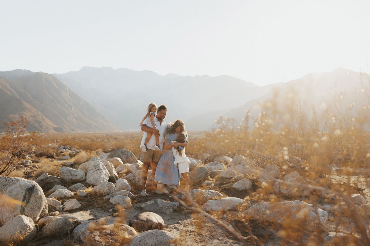 Family of four poses on rocky desert terrain with golden sunlight and mountain backdrop.