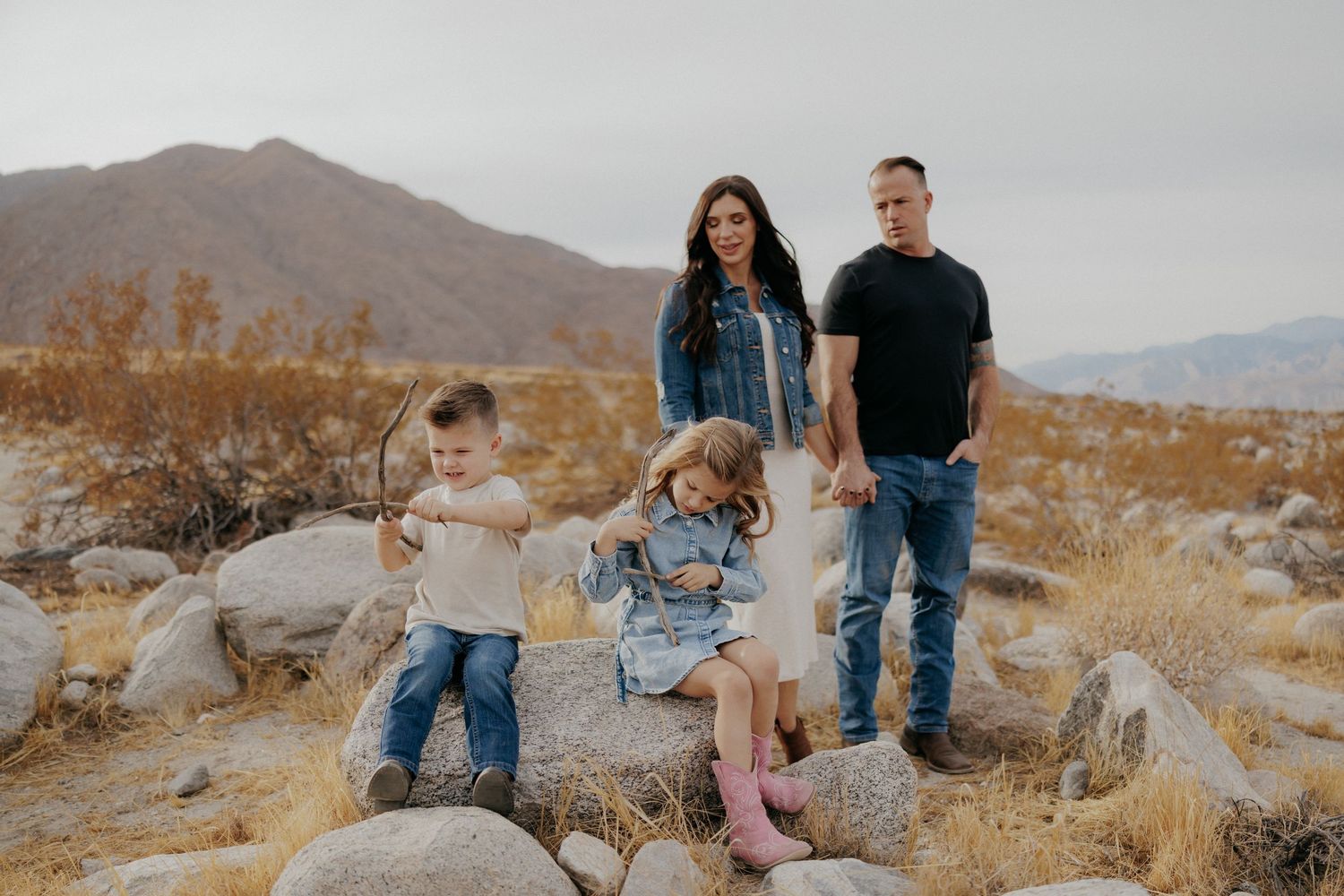 Family of four posing outdoors in desert landscape with mountains, two children playing on rocks while parents stand behind them.
