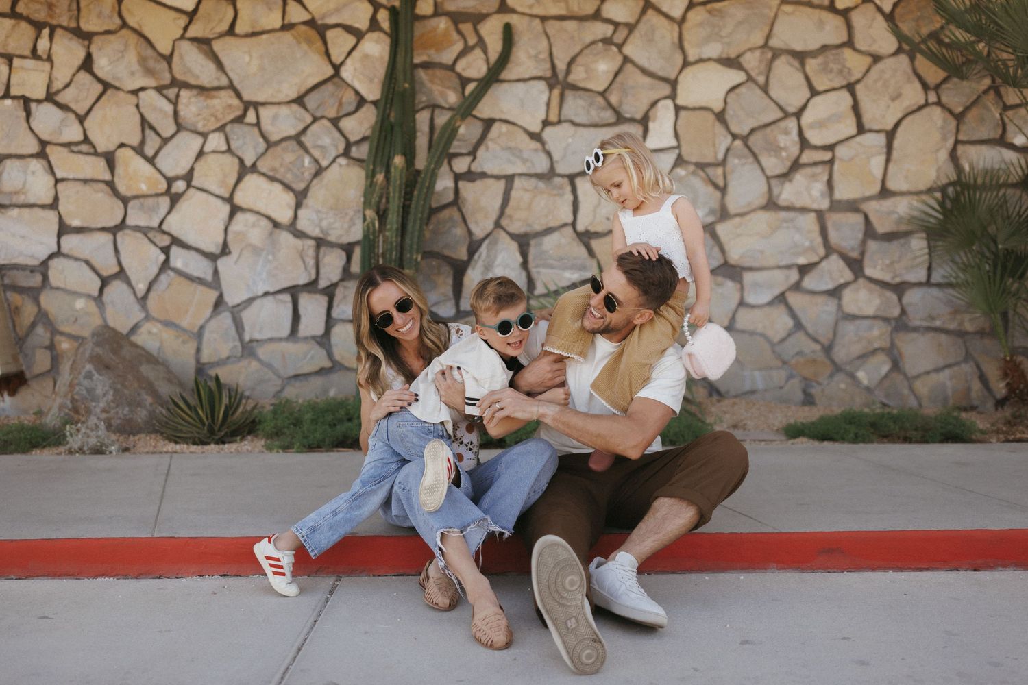 Family of four sitting against a stone wall outdoors, parents and two kids smiling and looking at a phone.