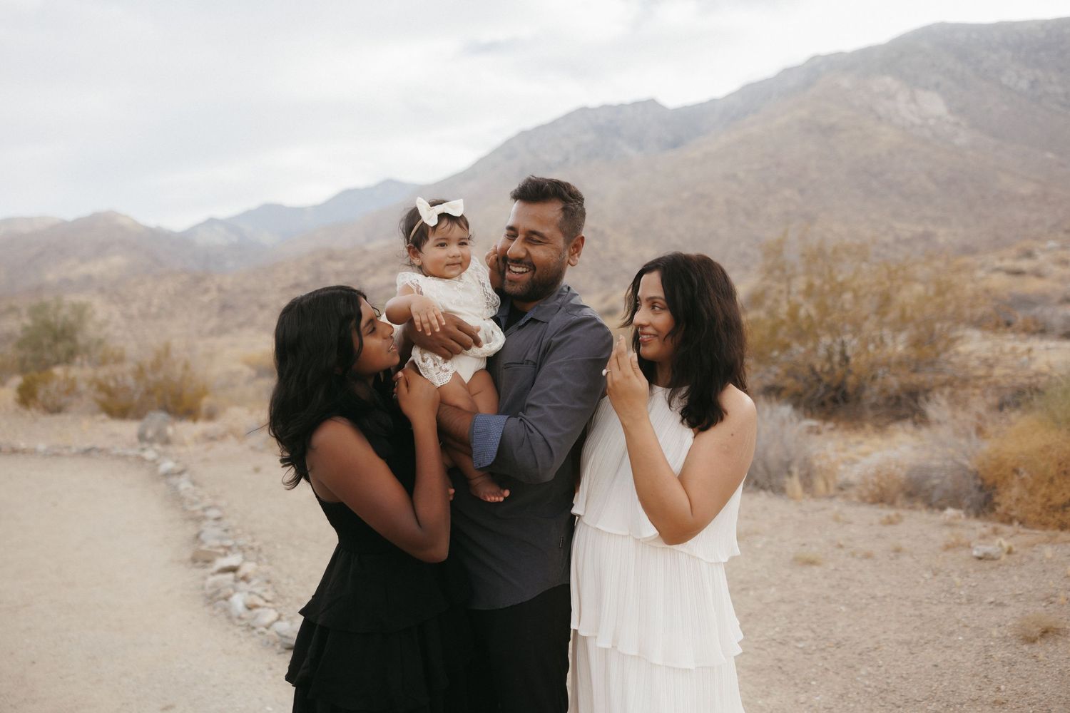 Family of three poses outdoors in desert landscape with mountains, parents holding baby girl in white dress.