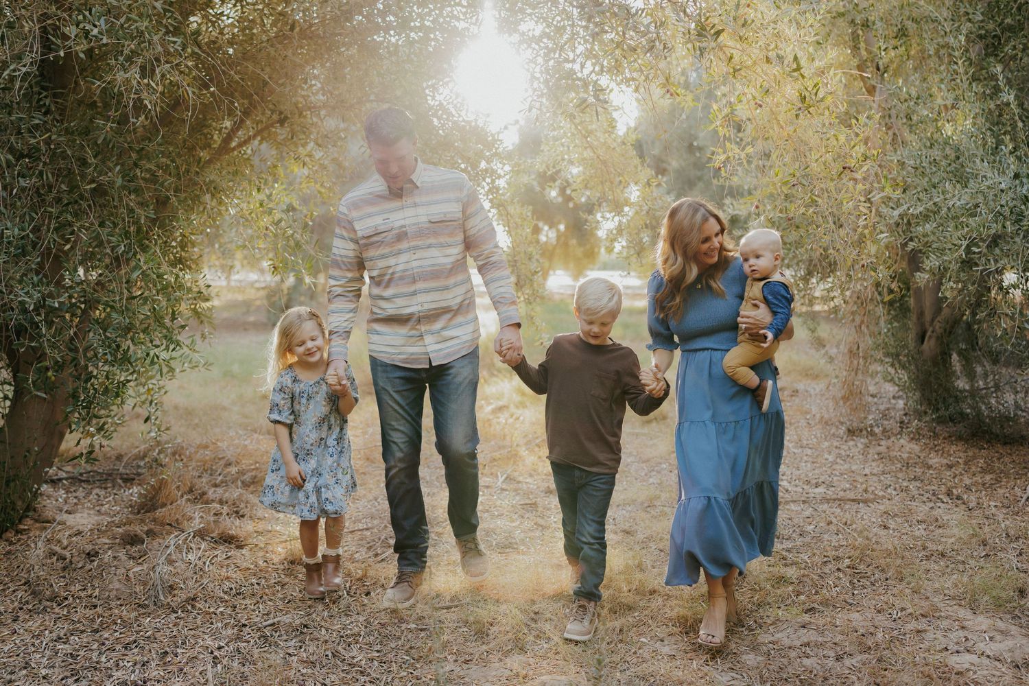 Family of five walking hand-in-hand through a sunlit olive grove, parents with three young children on a dirt path.