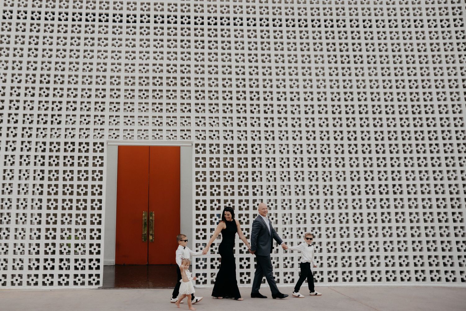 Family walking past a decorative white lattice wall with a red door in a modern architectural setting.