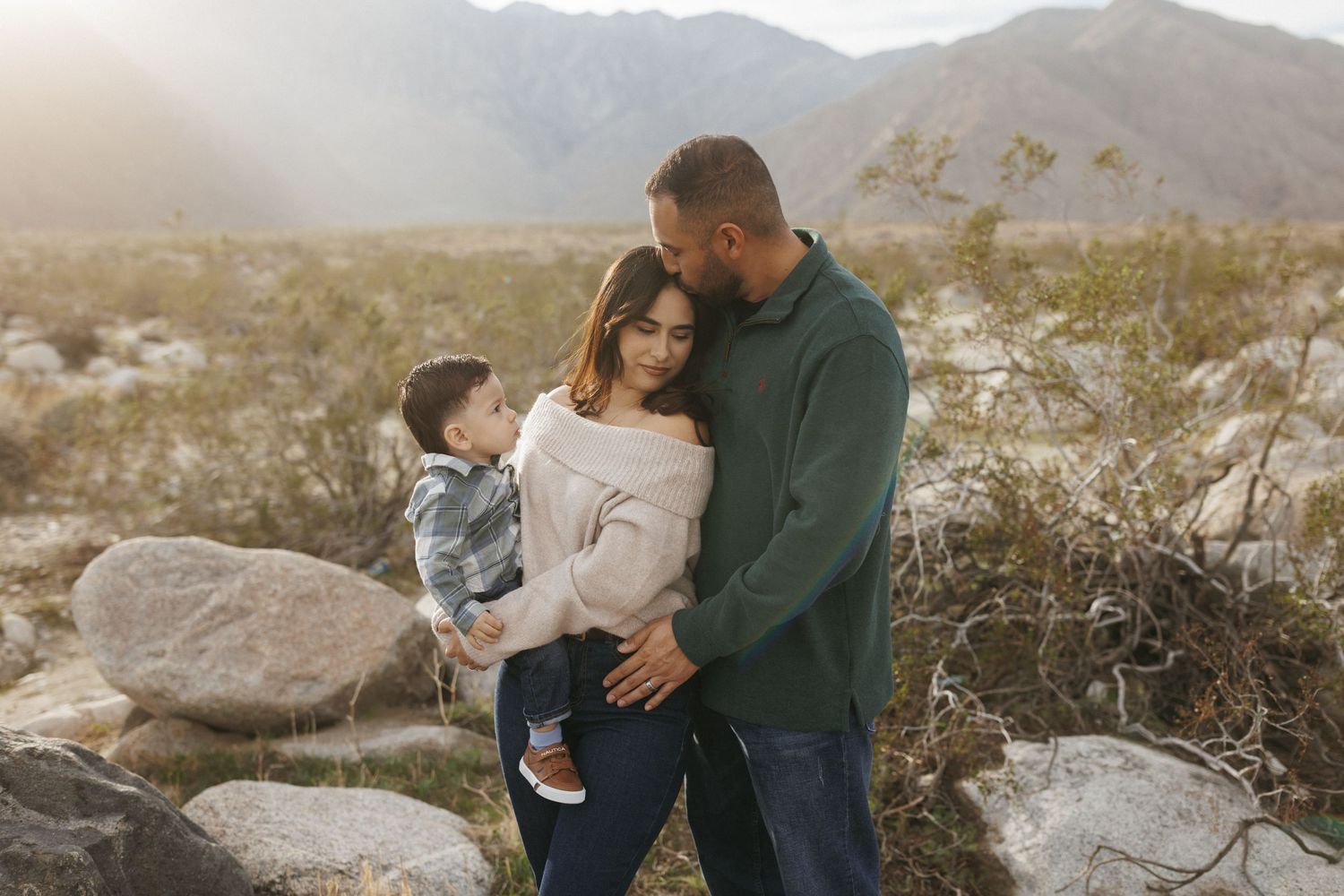 Family of three poses in desert landscape with mountains and golden sunlight behind them.