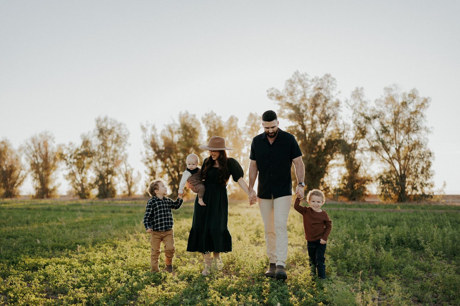 Family of five walking through a sunny green field with trees in the background during golden hour.