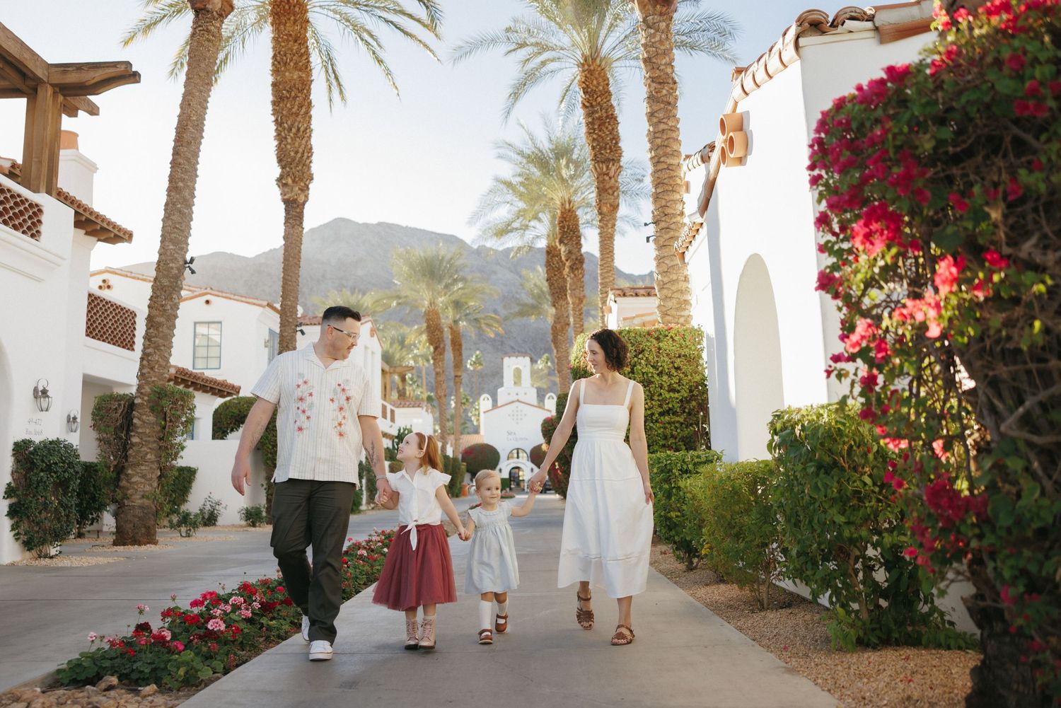 Family of four walking hand-in-hand along a sunny palm-lined resort path with white buildings and blooming bougainvillea.