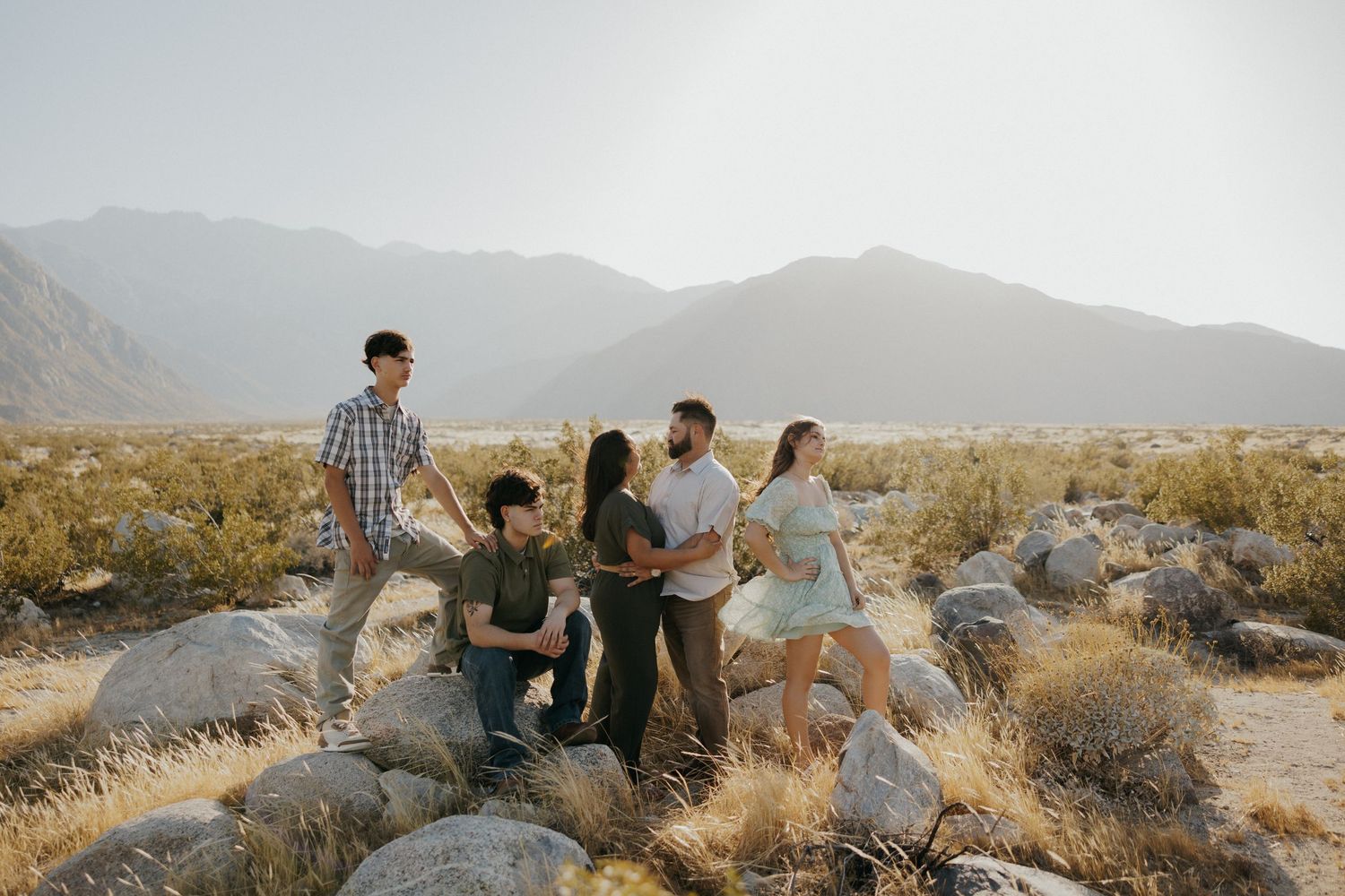 Five friends gather on rocky boulders in a sunny desert landscape with mountains in the background.