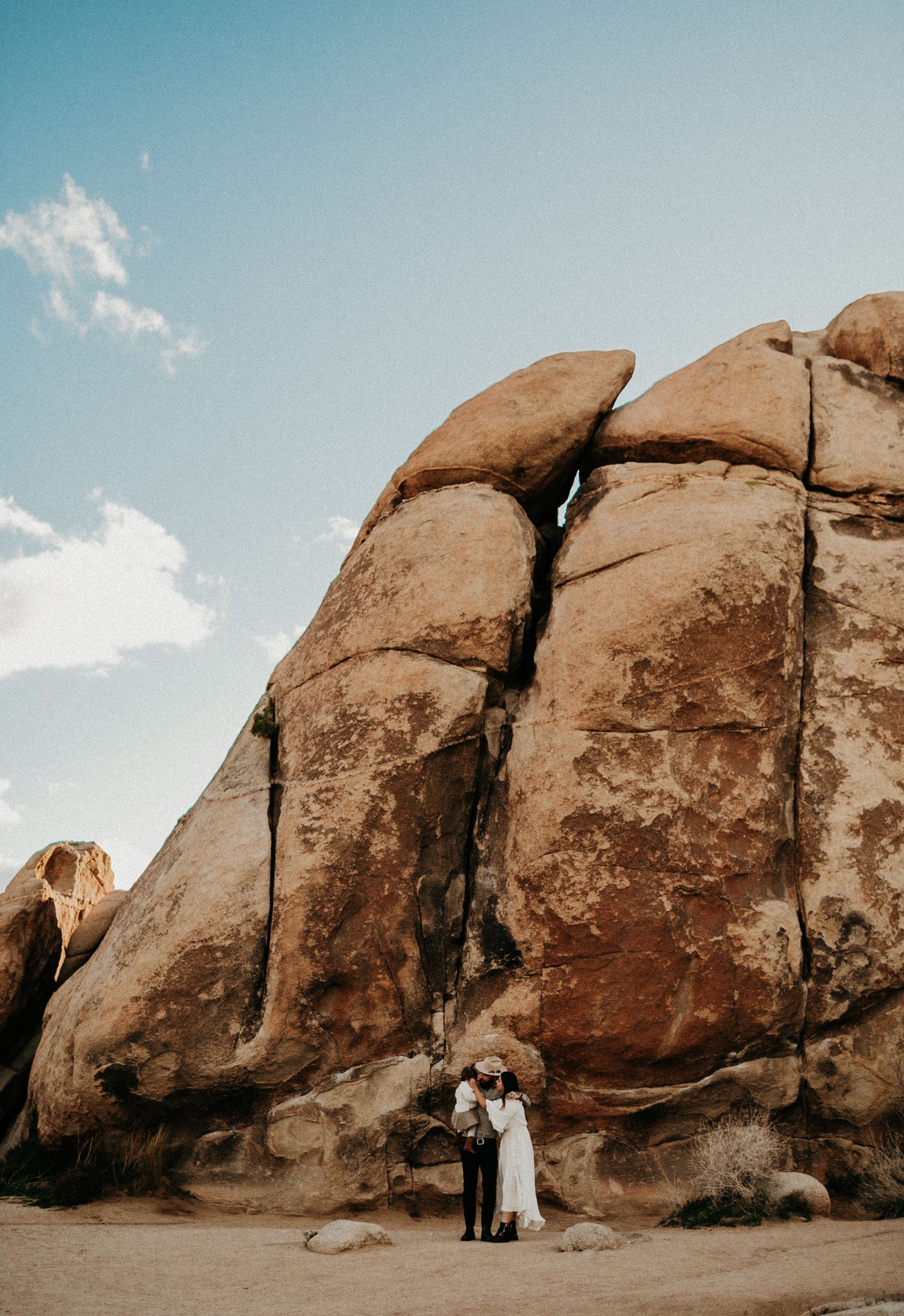 Couple standing before massive desert rock formations in Joshua Tree under a blue sky.