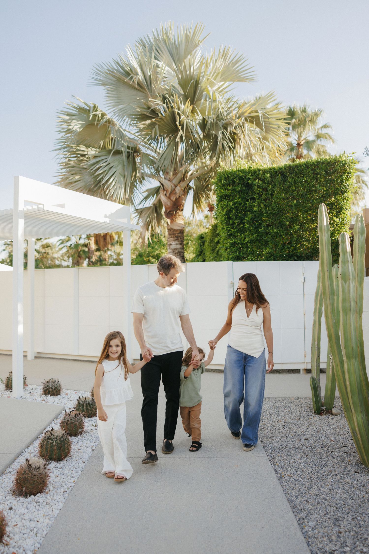 A happy family of four walks hand-in-hand along a sunny path surrounded by palm trees and cacti.