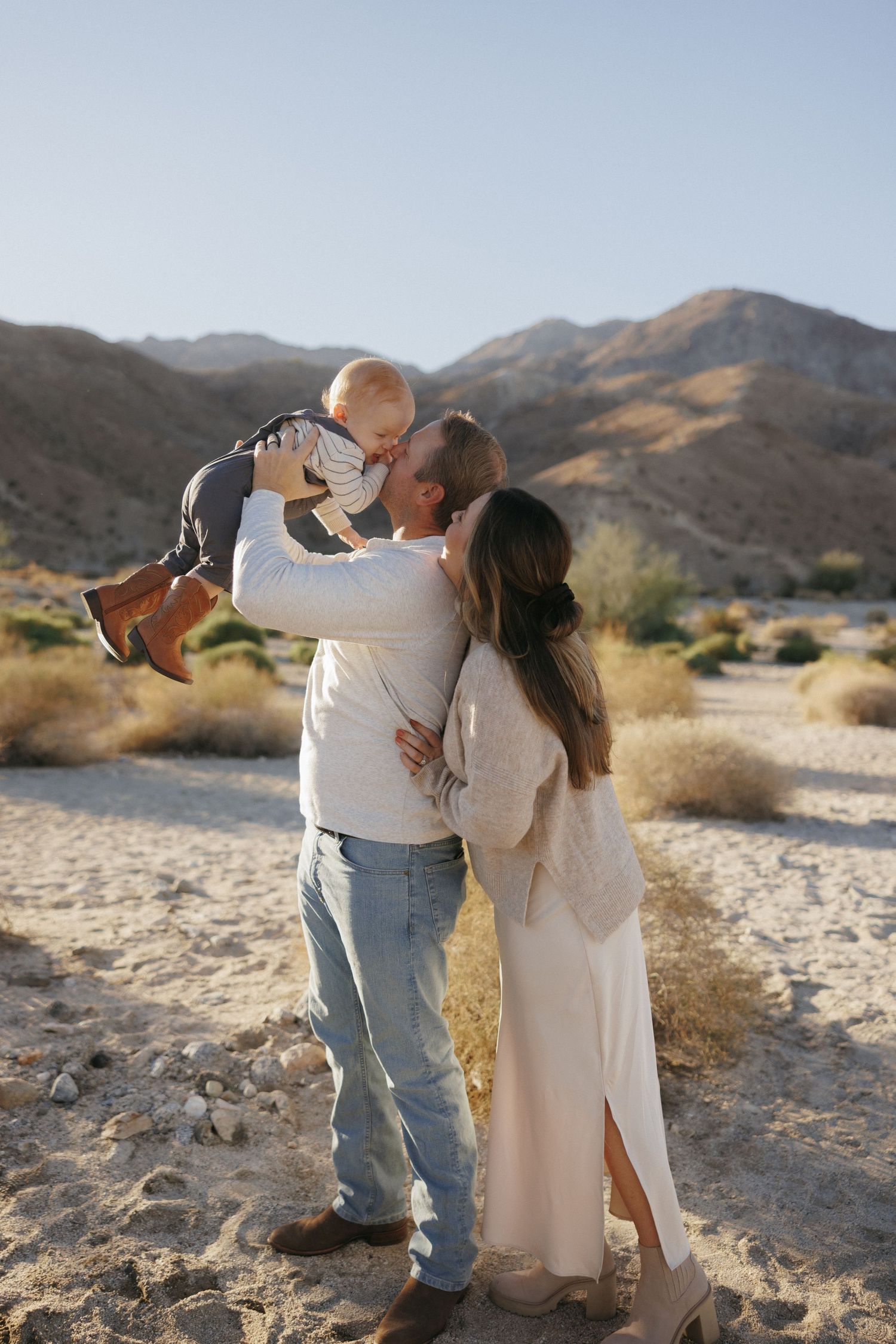 A couple kisses their baby in a sunny desert landscape with mountains in the background.