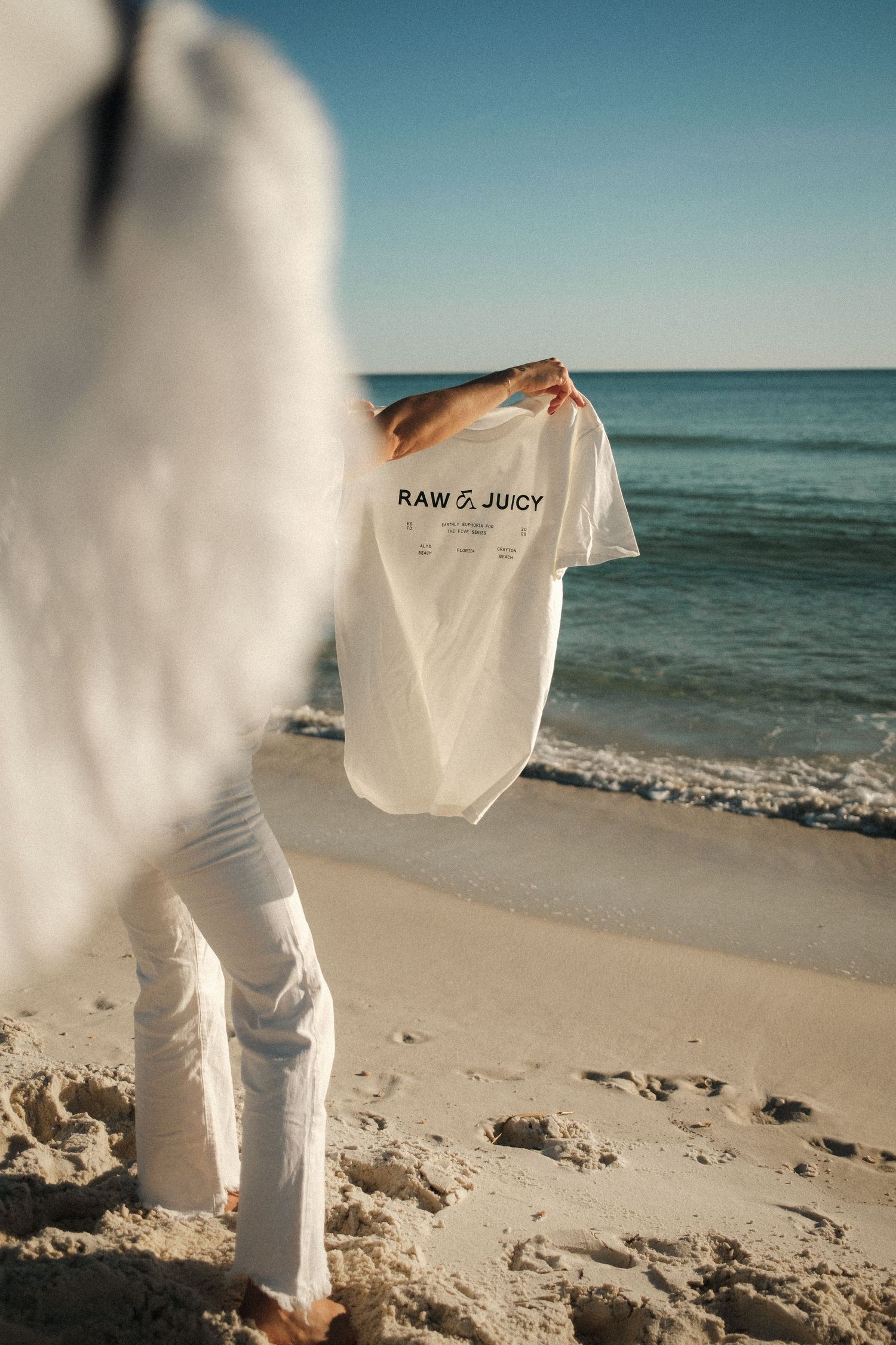 White t-shirt held up against ocean backdrop on sunny beach.
