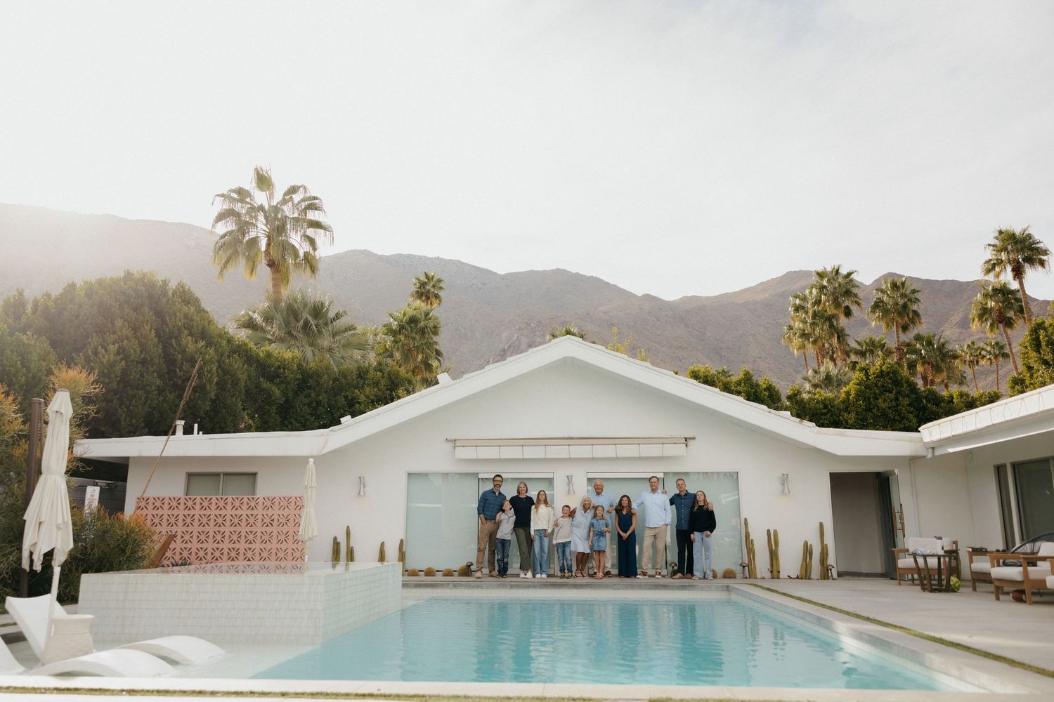 Group of people posing by a pool at a white mid-century modern home with palm trees and mountains in Palm Springs.