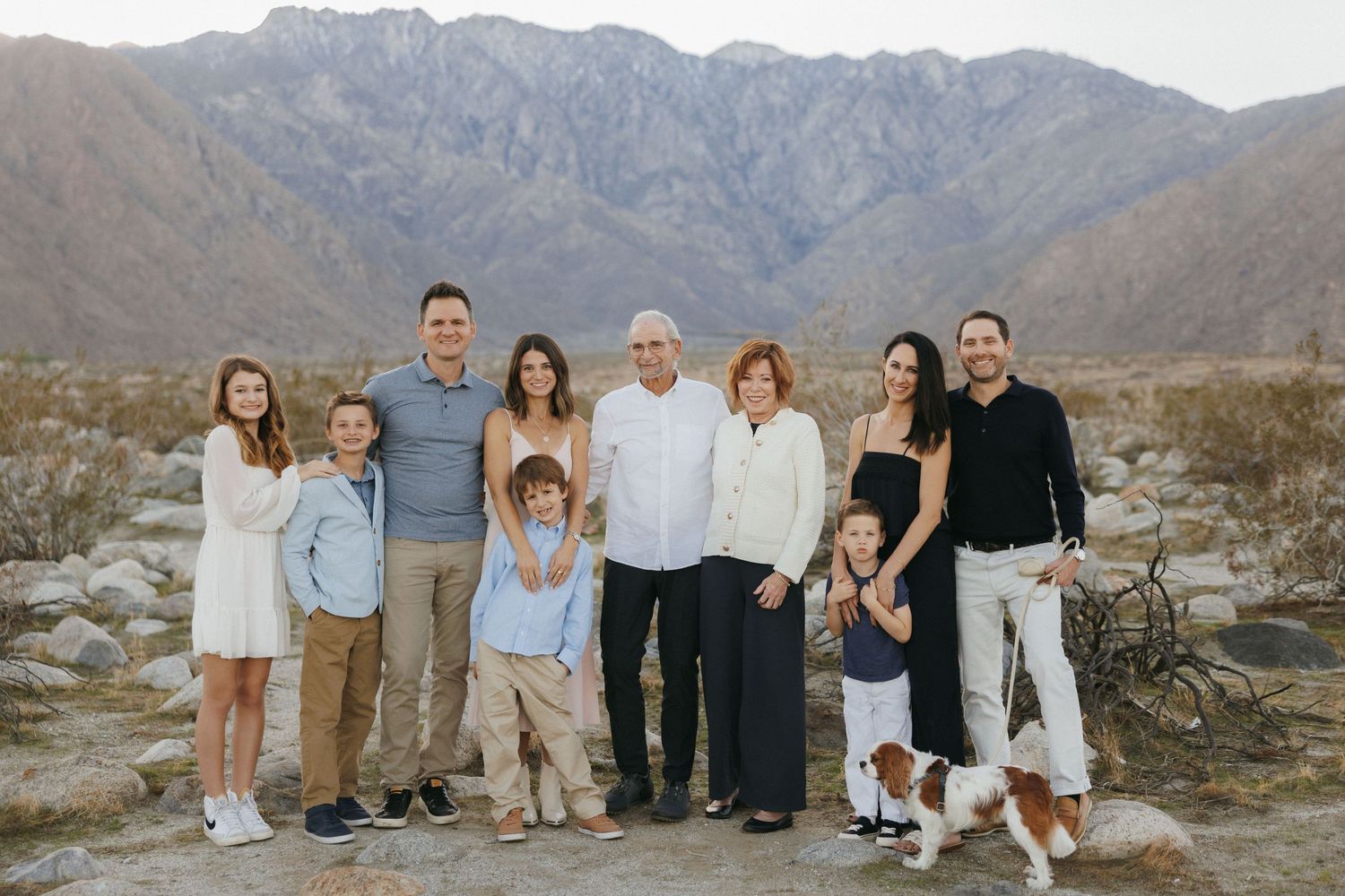 Multi-generational family group portrait outdoors with desert mountains backdrop, including children and a dog.