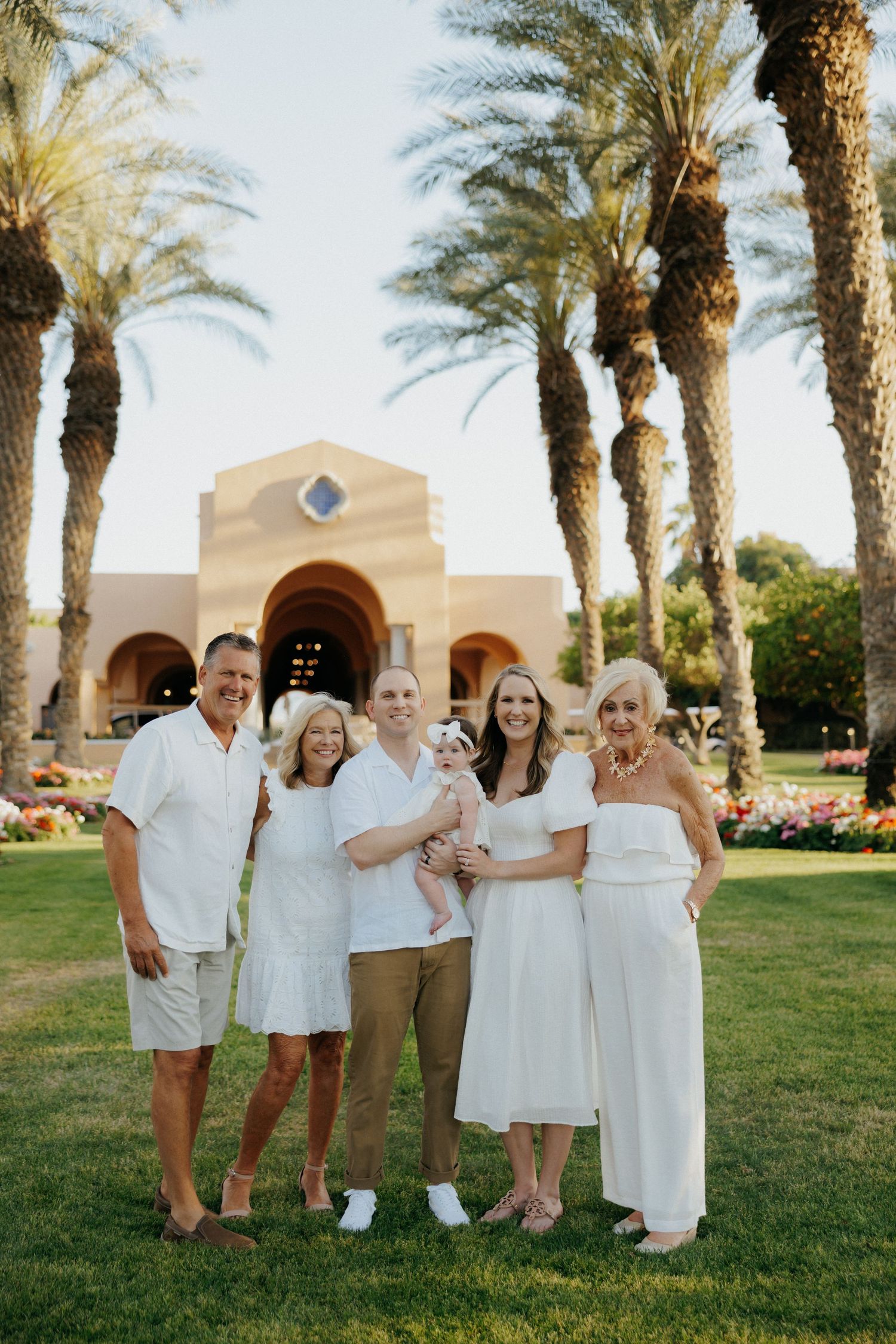 Family of six posing in white outfits on green lawn with tall palm trees and Mediterranean building backdrop.