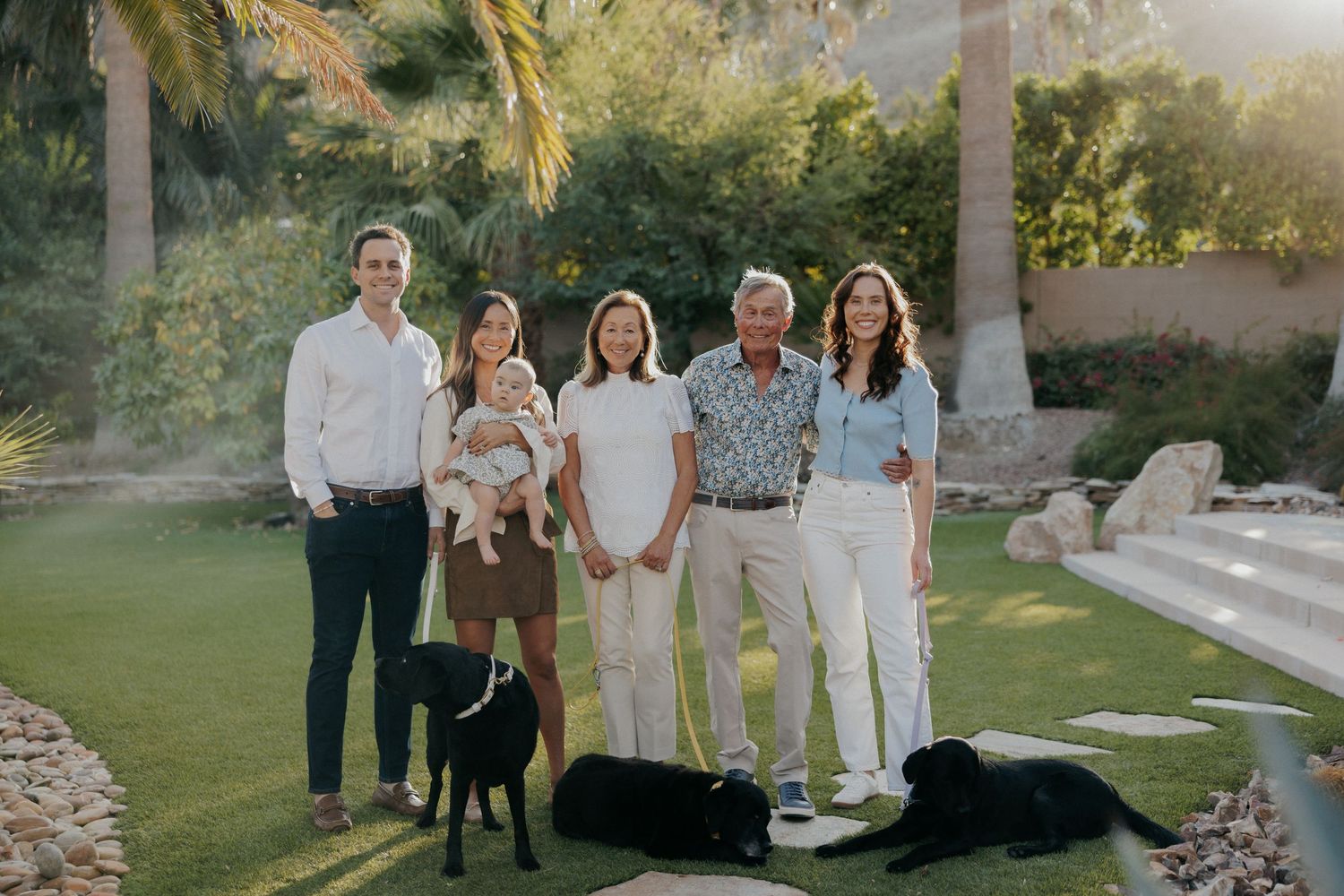 Family of five posing outdoors on green lawn with two black dogs, palm trees, and warm golden sunlight in background.