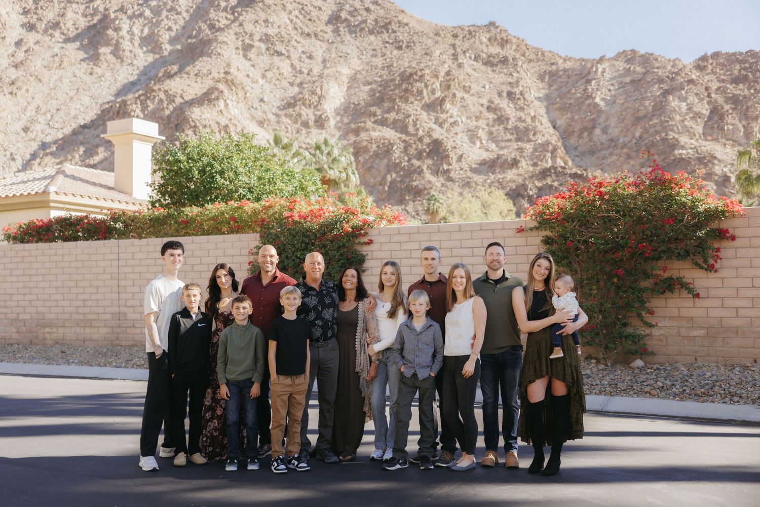 Large family group posing outdoors with desert mountains and red flowering bushes in sunny Palm Springs backdrop.