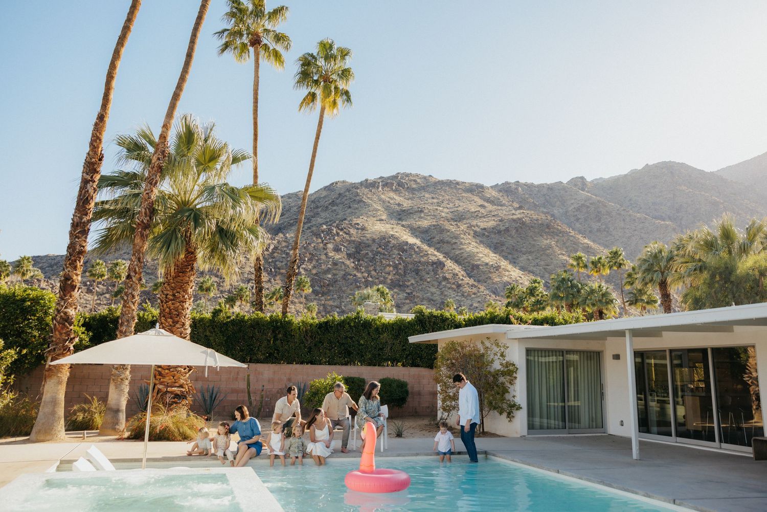 Group of people enjoying a poolside gathering at a mid-century modern home with palm trees and mountains in Palm Springs.