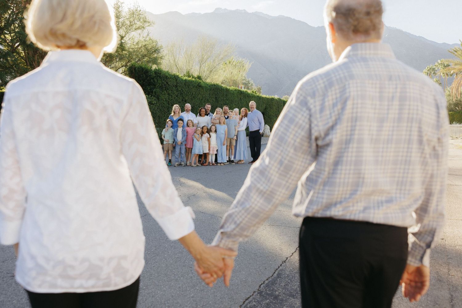 Elderly couple holding hands walking toward large family group on road with mountain backdrop.